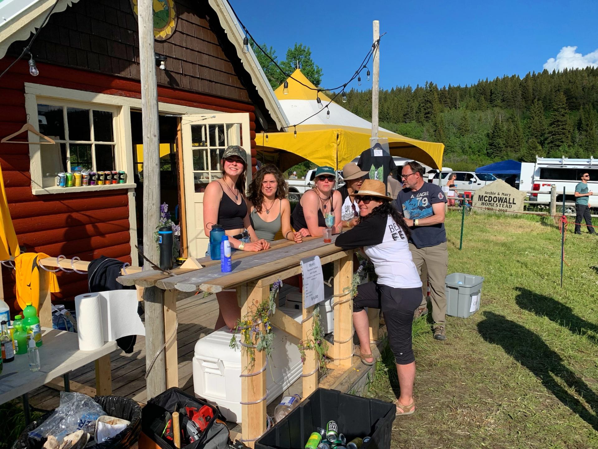 People stand at a rustic outdoor bar in front of a red cabin, with yellow tents, a grassy field, and forested hills under a blue sky.
