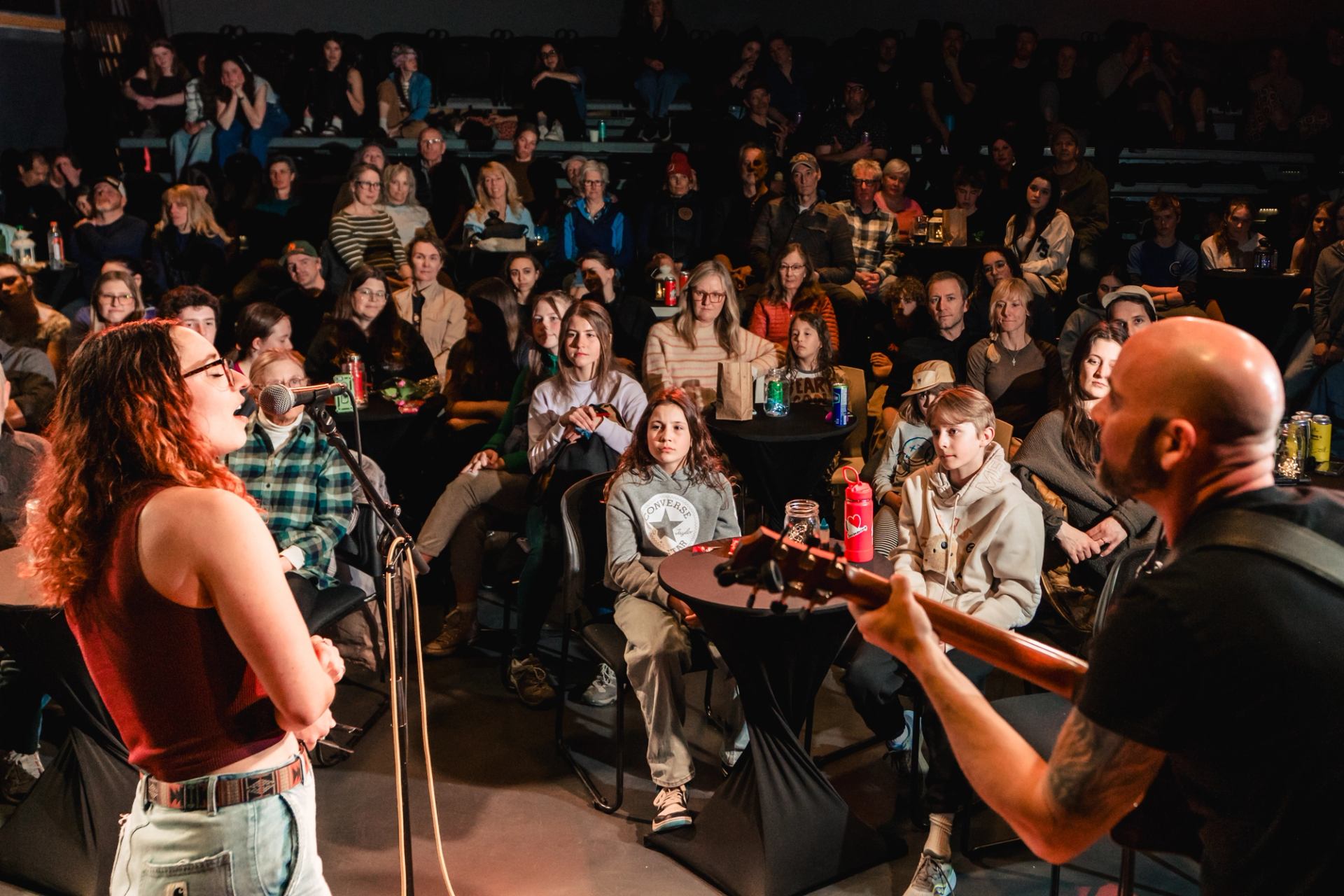 Musician sings into microphone as guitarist plays on stage before seated audience.
