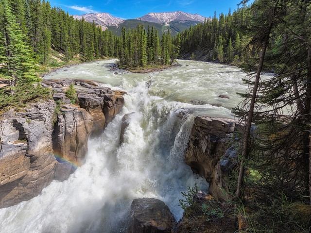 Wide waterfall rushing through rocky gorge, surrounded by dense evergreens and distant mountains.