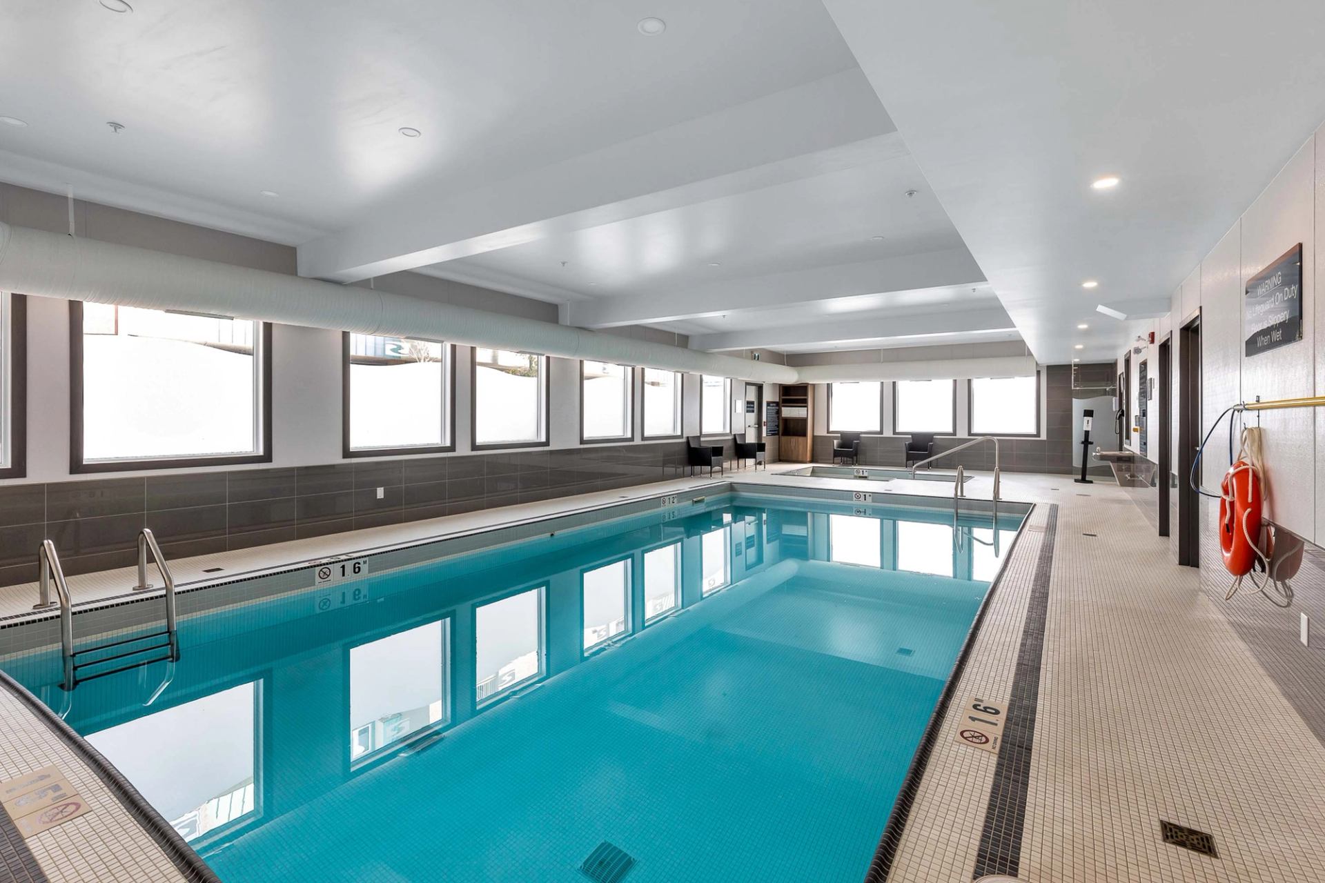 Indoor swimming pool with tiled deck, windows along one wall and safety equipment mounted.