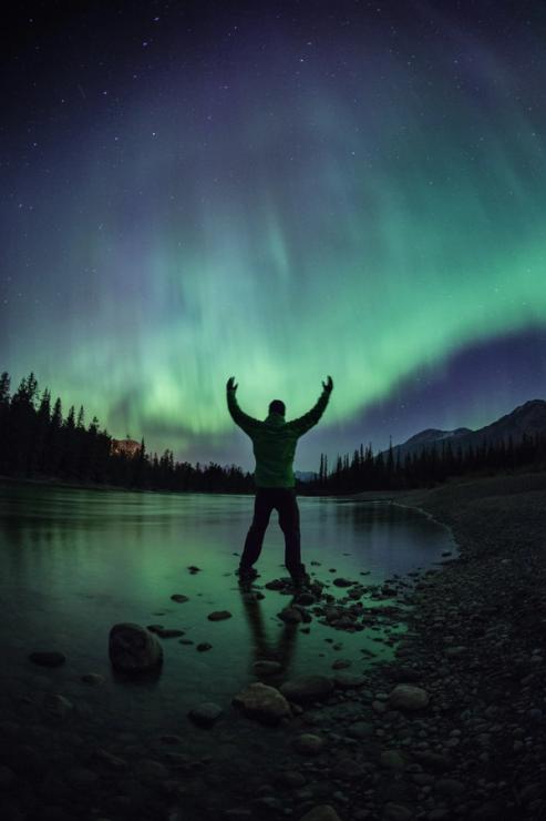 Person standing near a lake with arms reaching up to the northern lights in the Jasper National Park Dark Sky Preserve.