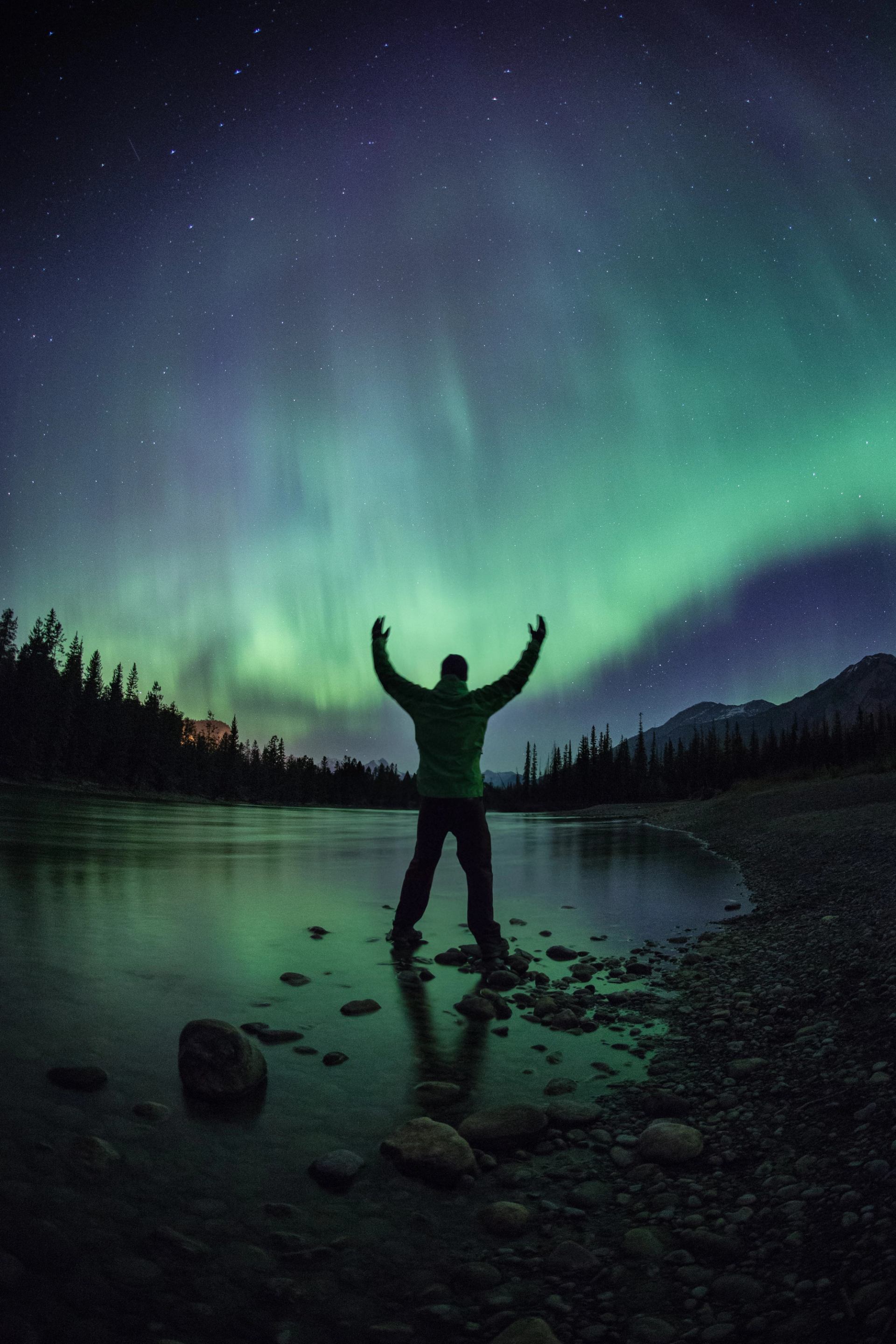 Person with arms raised near lake under northern lights, Jasper Dark Sky Preserve.