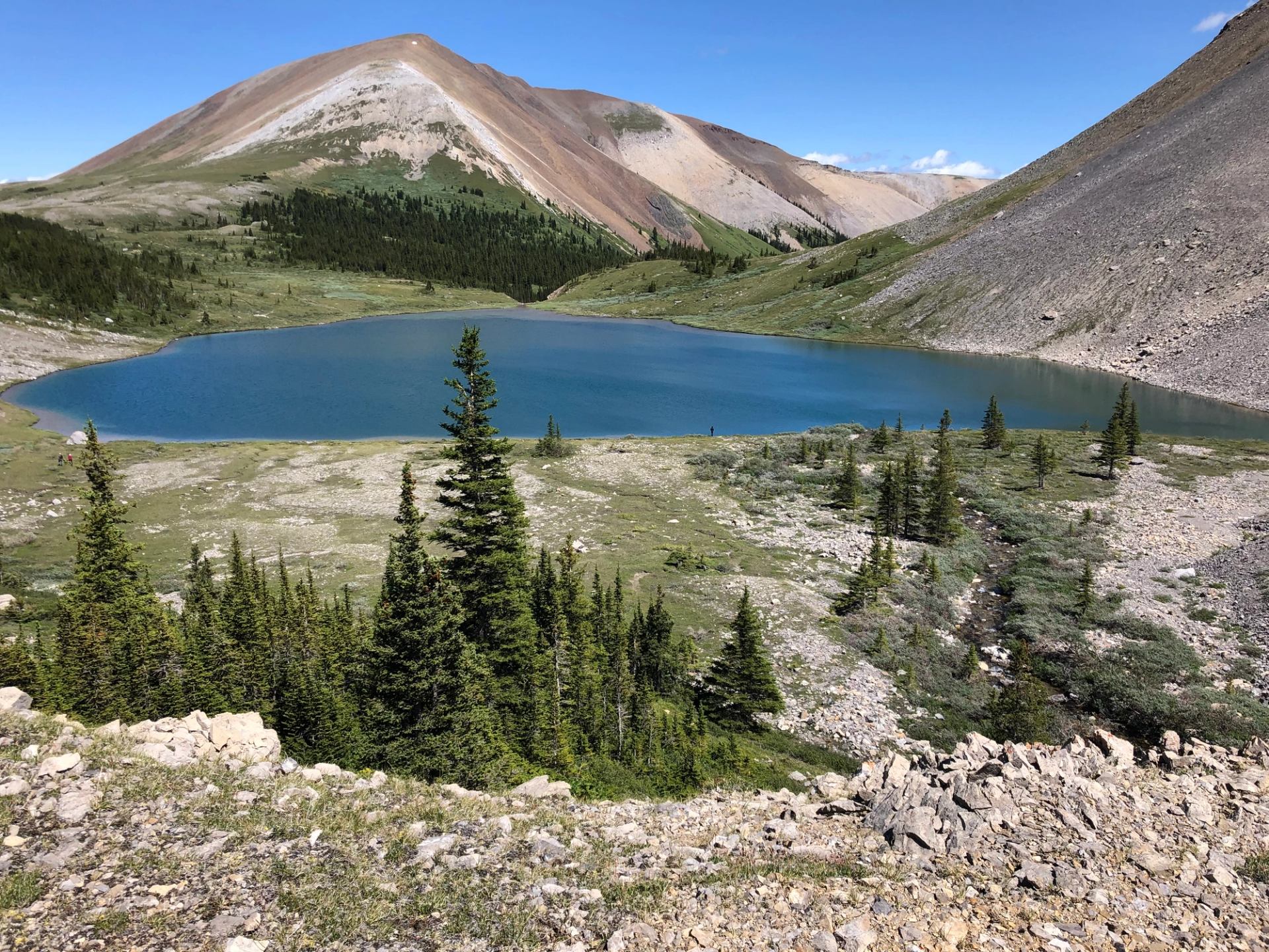 A turquoise alpine lake surrounded by rocky slopes, evergreen trees, and rolling mountain terrain under a clear blue sky.