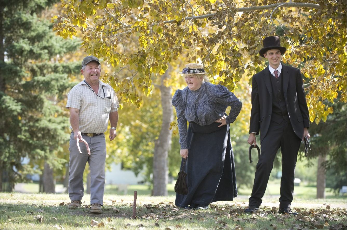 Three people playing horseshoes outdoors under fall foliage in a park-like setting.