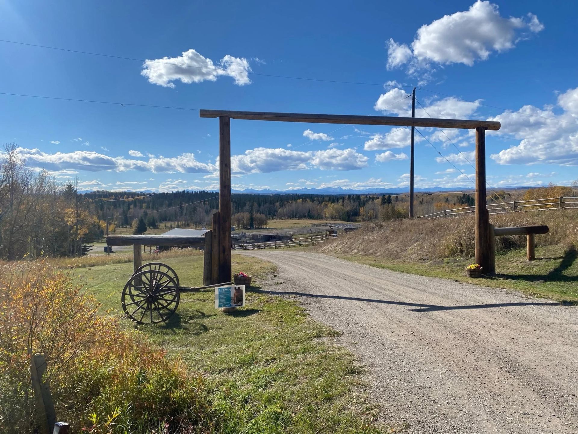 Rural gravel road passing under wooden ranch gate with wagon wheel and distant trees.