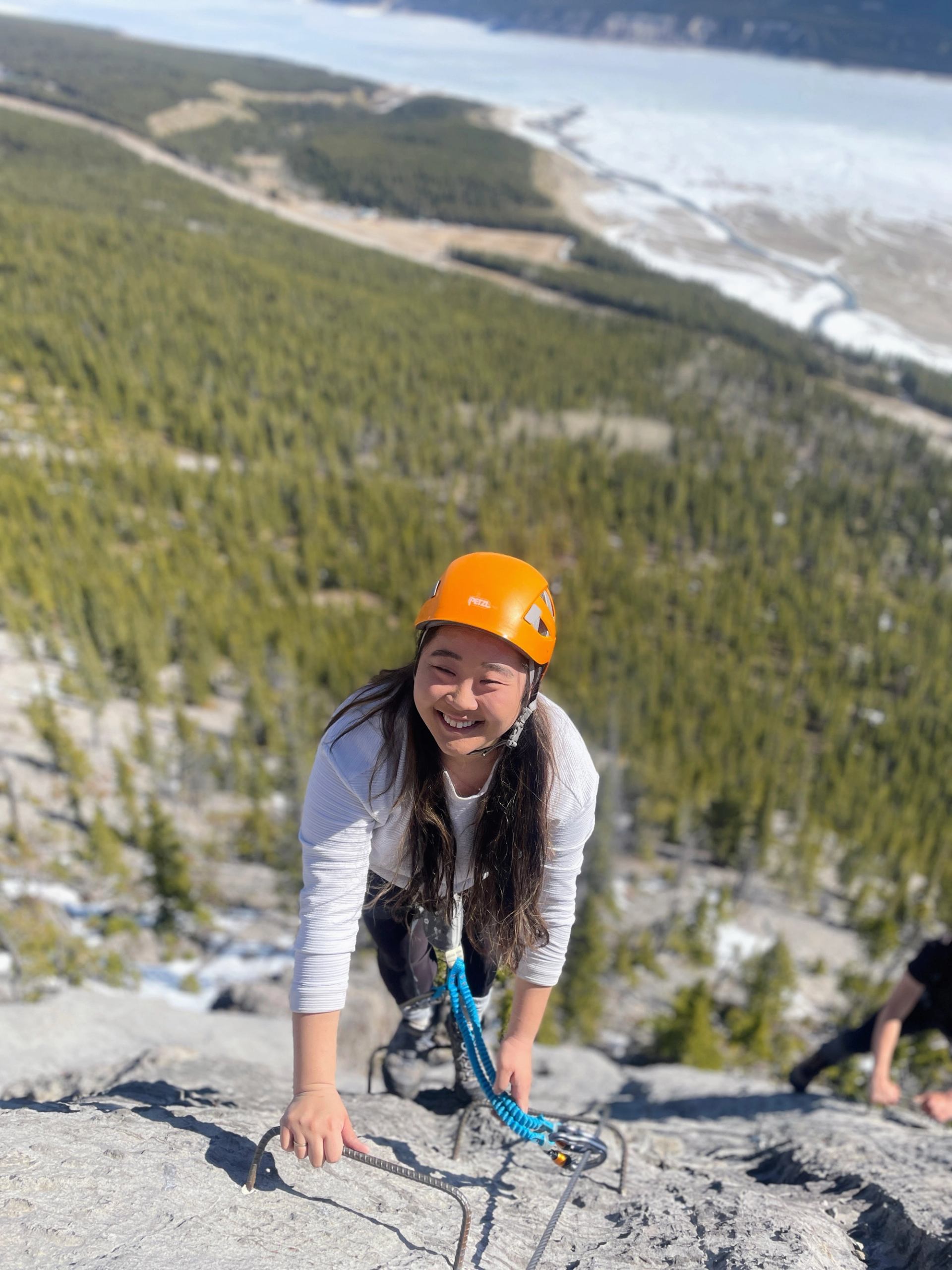 A smiling woman in an orange helmet ascends a via ferrata with a mountainous landscape and river in the background.