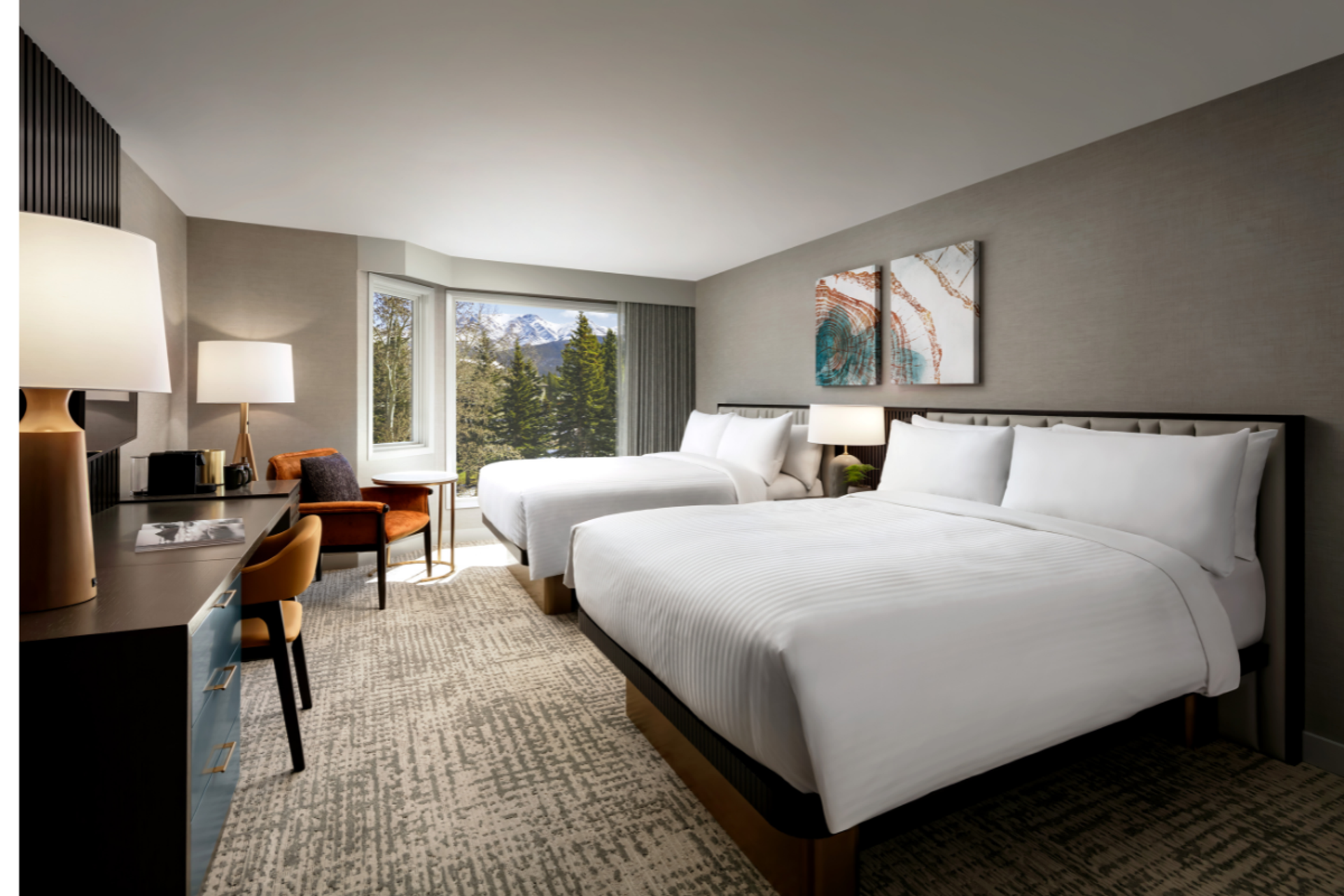 Hotel room with two beds, desk, armchair and window overlooking forested mountains.
