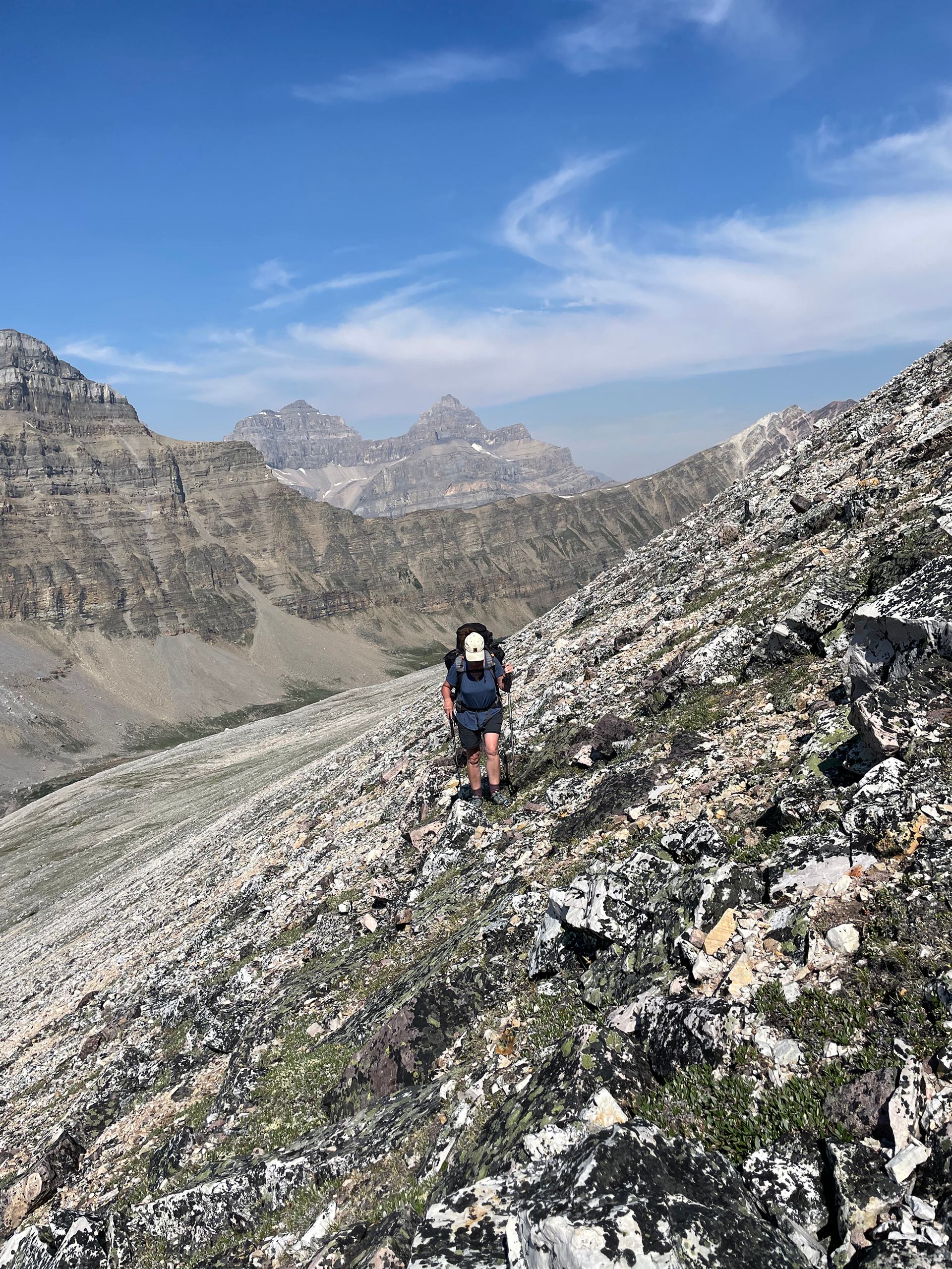 Hiker with backpack climbs steep rocky mountain slope under clear blue sky and distant peaks.