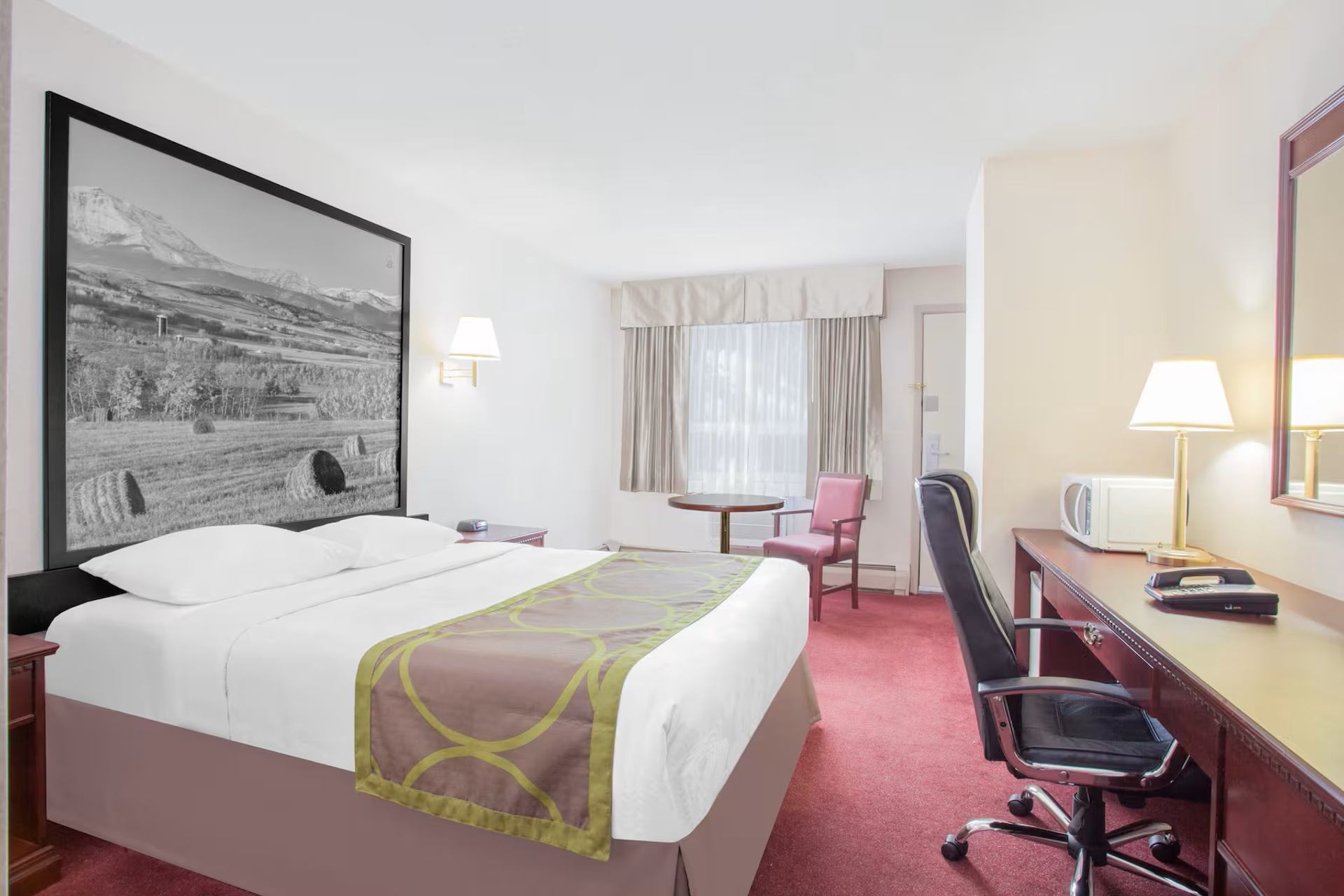 Hotel room with bed, landscape photograph, desk, chairs, microwave and window.