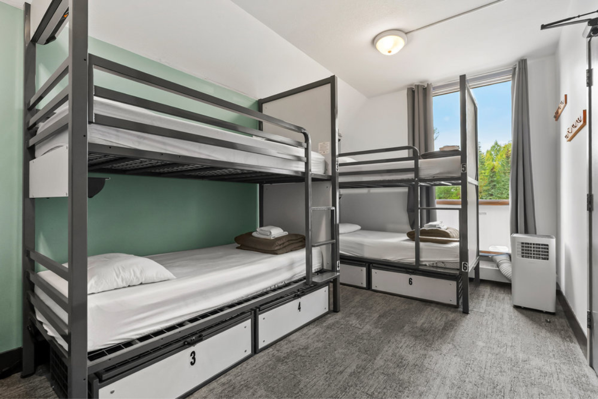 Four-bed dorm room with modern metal bunk beds, storage lockers, window with a view.