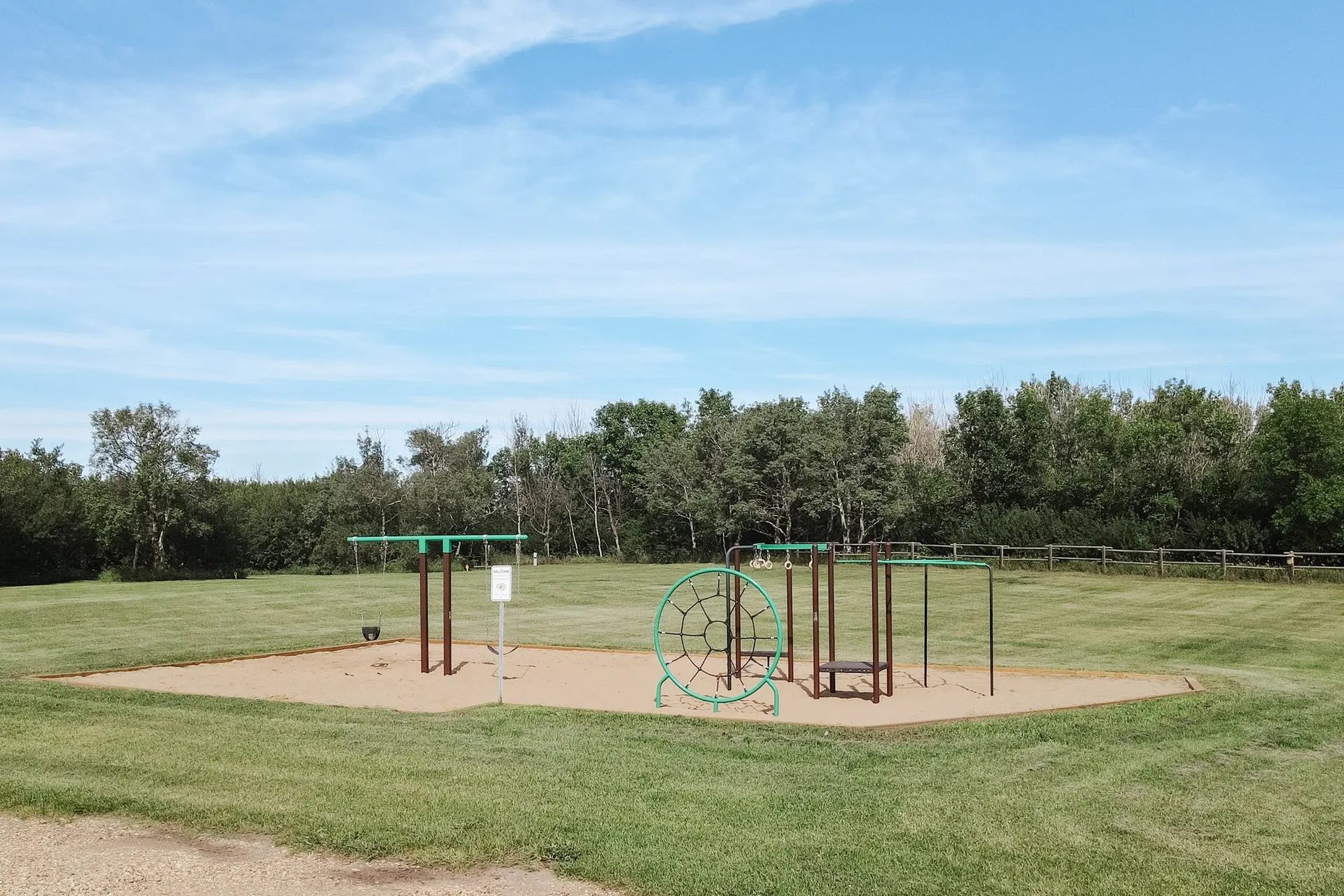 Playground with swings and climbing ring on sand, surrounded by grass and trees.