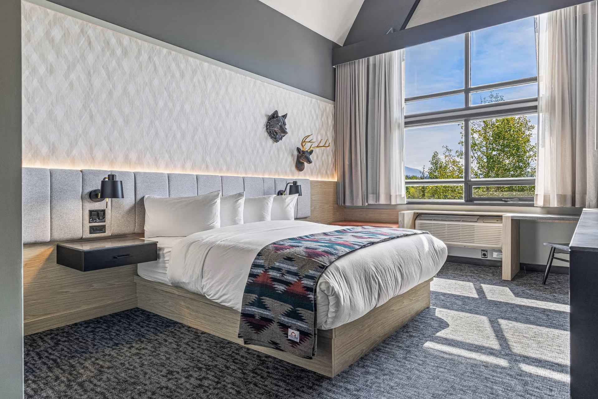 Modern hotel room with a king bed, patterned throw, wood accents, and large window with tree views.