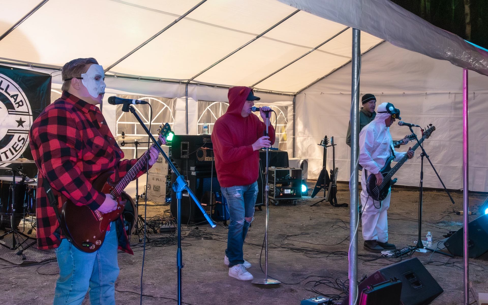 Rock band in costumes performs on outdoor tent stage with guitars, microphones and speakers.