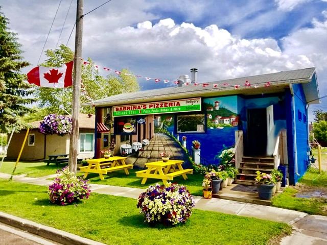 Colorful pizzeria exterior with mural, picnic tables, flowers, and Canadian flag.