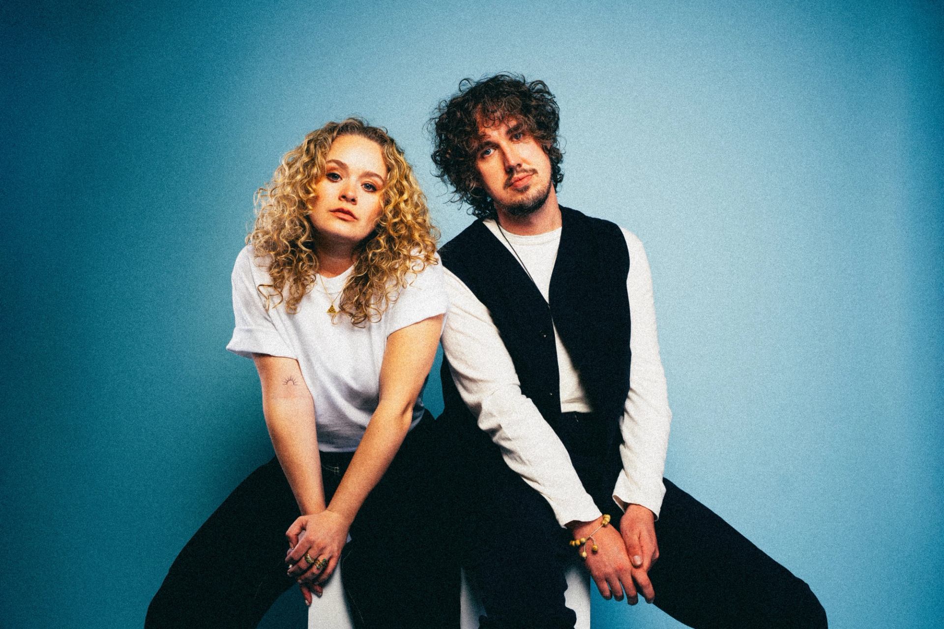 Two people sit side by side against a blue studio backdrop, facing forward.