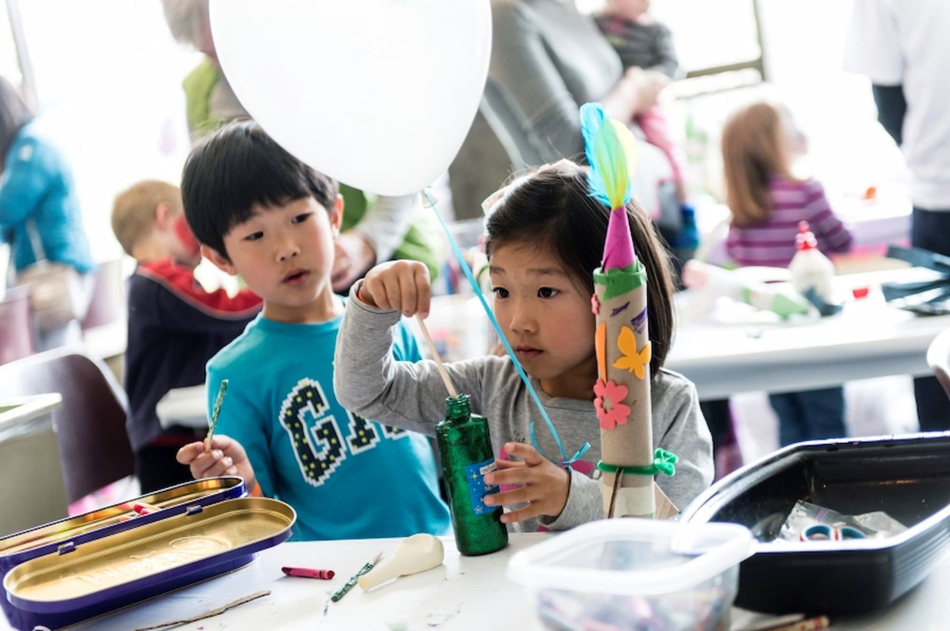 Children at a craft table decorate a cardboard rocket with paint, stickers and feathers.