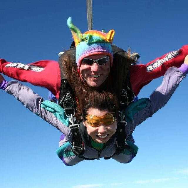 Skydivers wearing helmets and parachutes with arms extended against a clear blue sky.