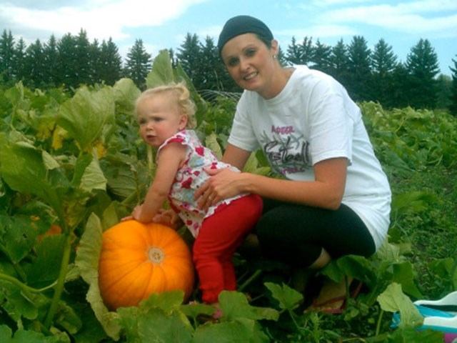 Child and adult in pumpkin patch with green plants and trees under blue sky.