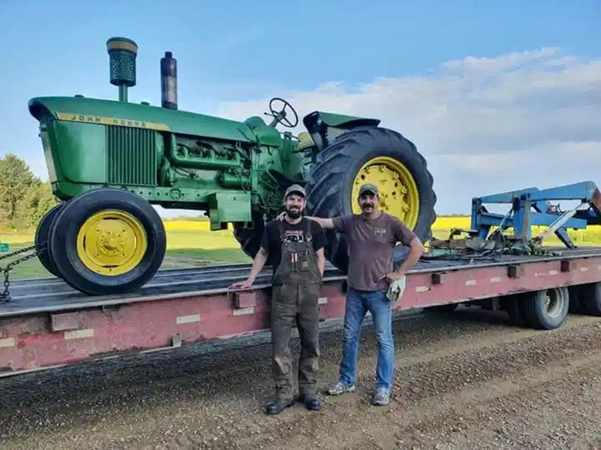 Two people stand on a flatbed trailer beside a large green John Deere tractor.