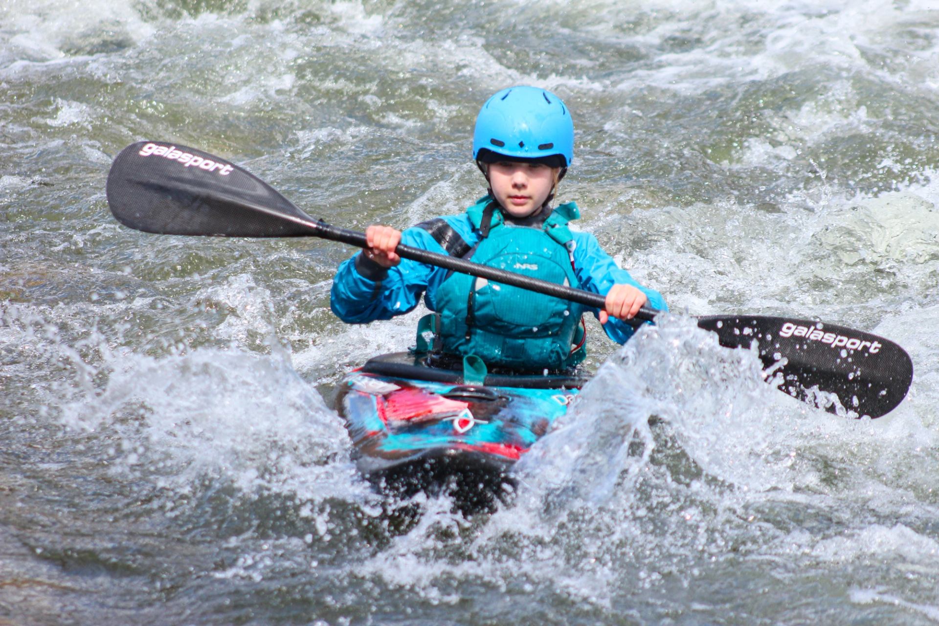 Kayaker paddling through swift rapids as water splashes around the kayak.