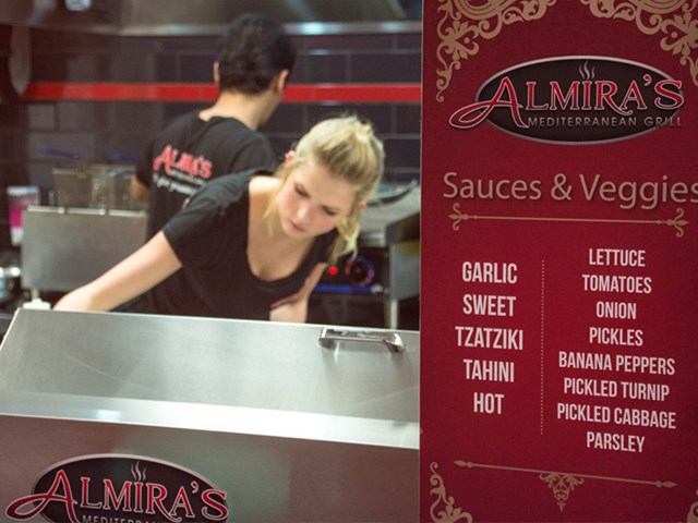 Kitchen staff at Almira’s Mediterranean Grill with a menu board listing sauces and veggies.