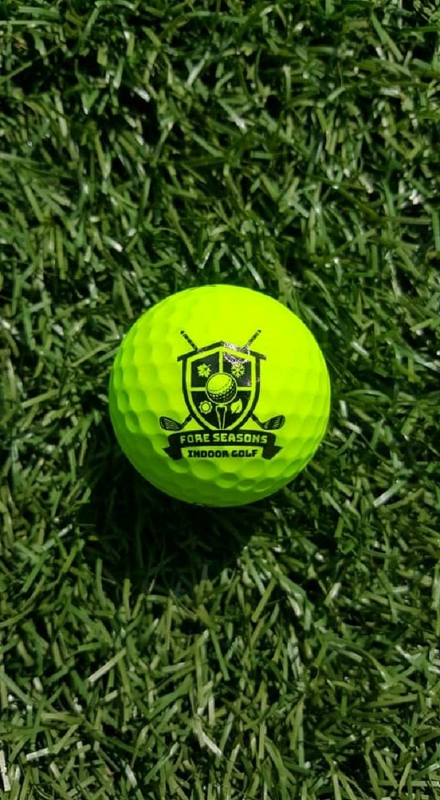 A bright yellow golf ball with the Fore Seasons Indoor Golf logo on artificial turf.