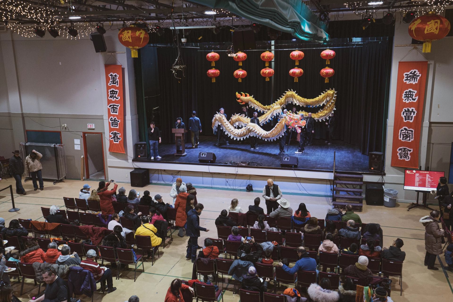 Stage performance of Chinese New Year dragon dance with red lanterns and seated audience.