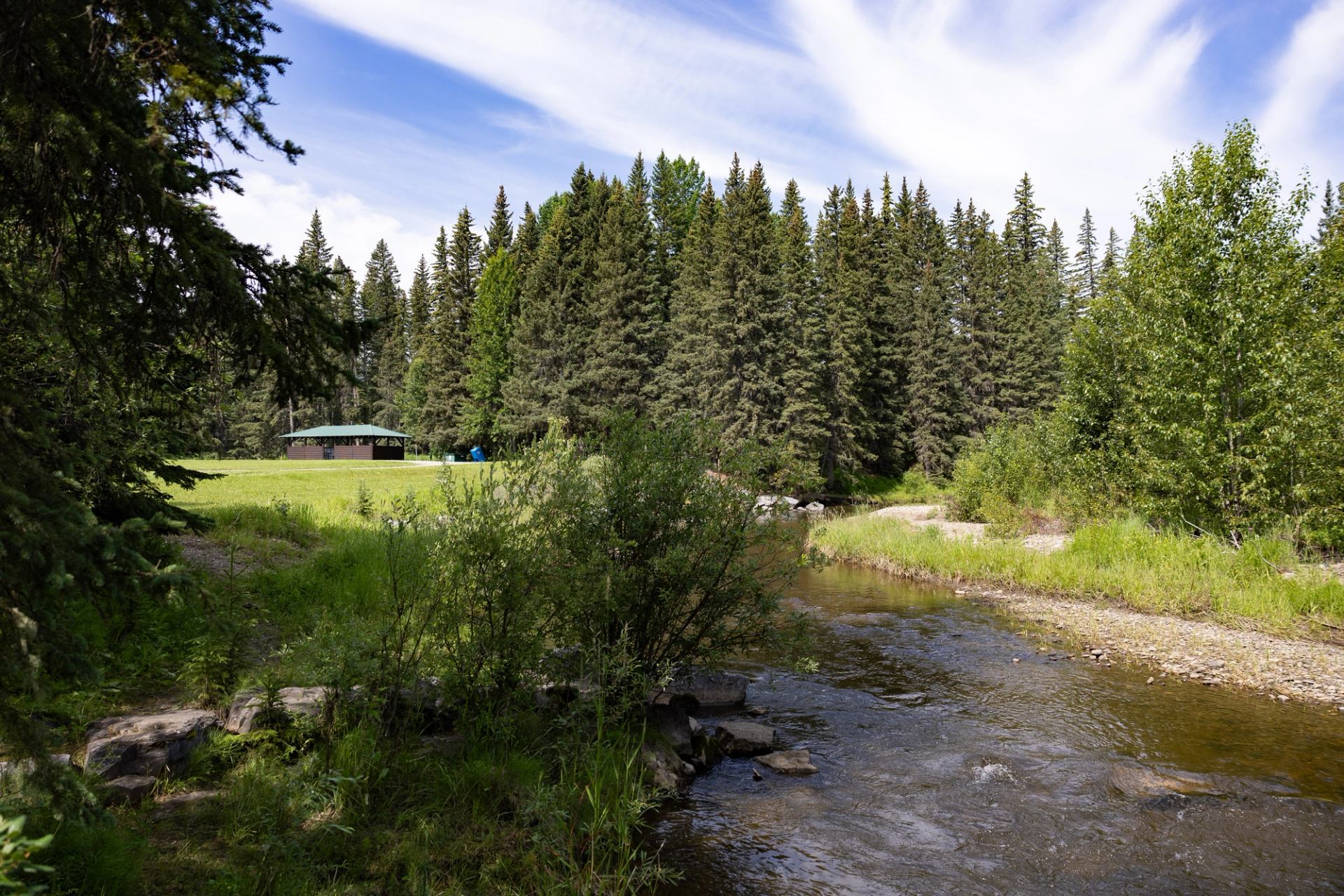 Shallow creek flows past grassy bank toward forest and small shelter building.
