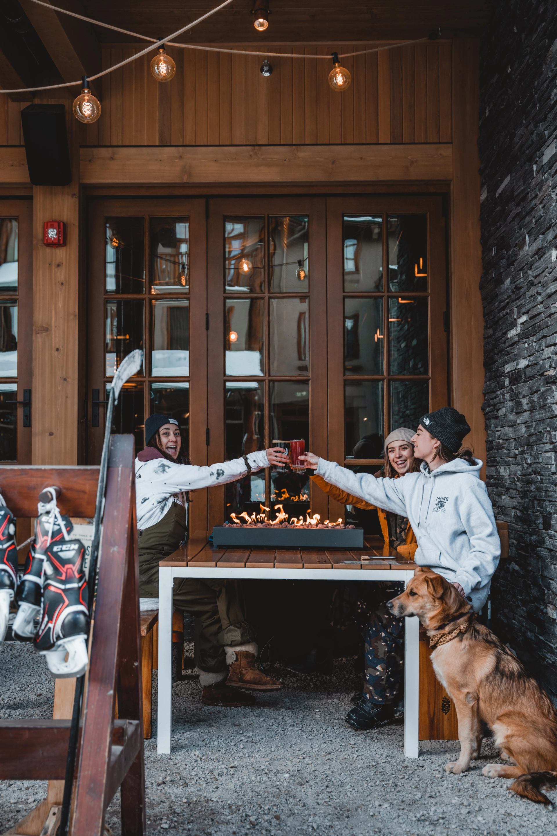 Three people and a dog toast drinks around a tabletop fire pit on a cozy outdoor patio with winter sports gear nearby.