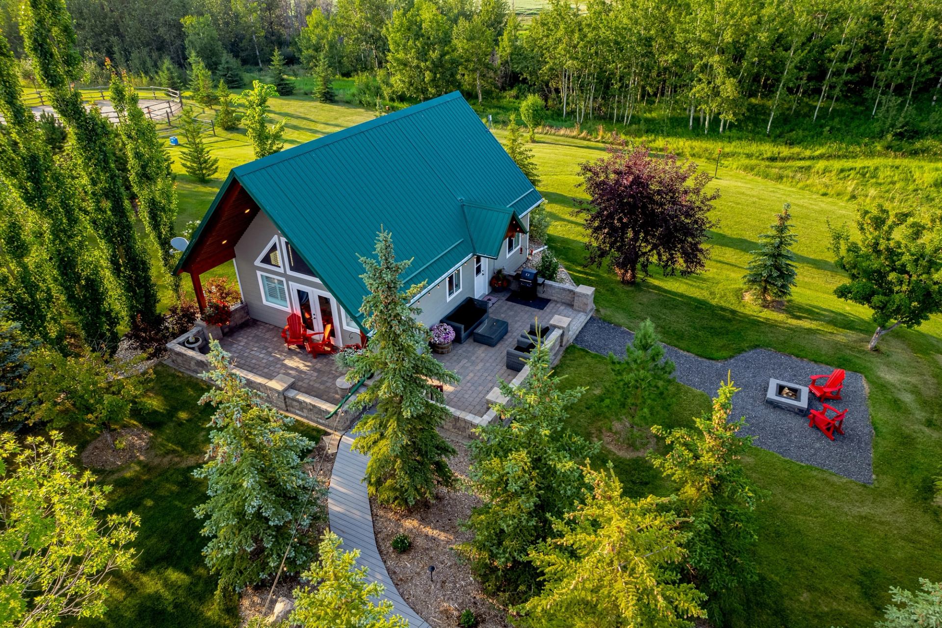 A cozy cabin with green roof surrounded by trees, patio seating, and a firepit in a lush yard.
