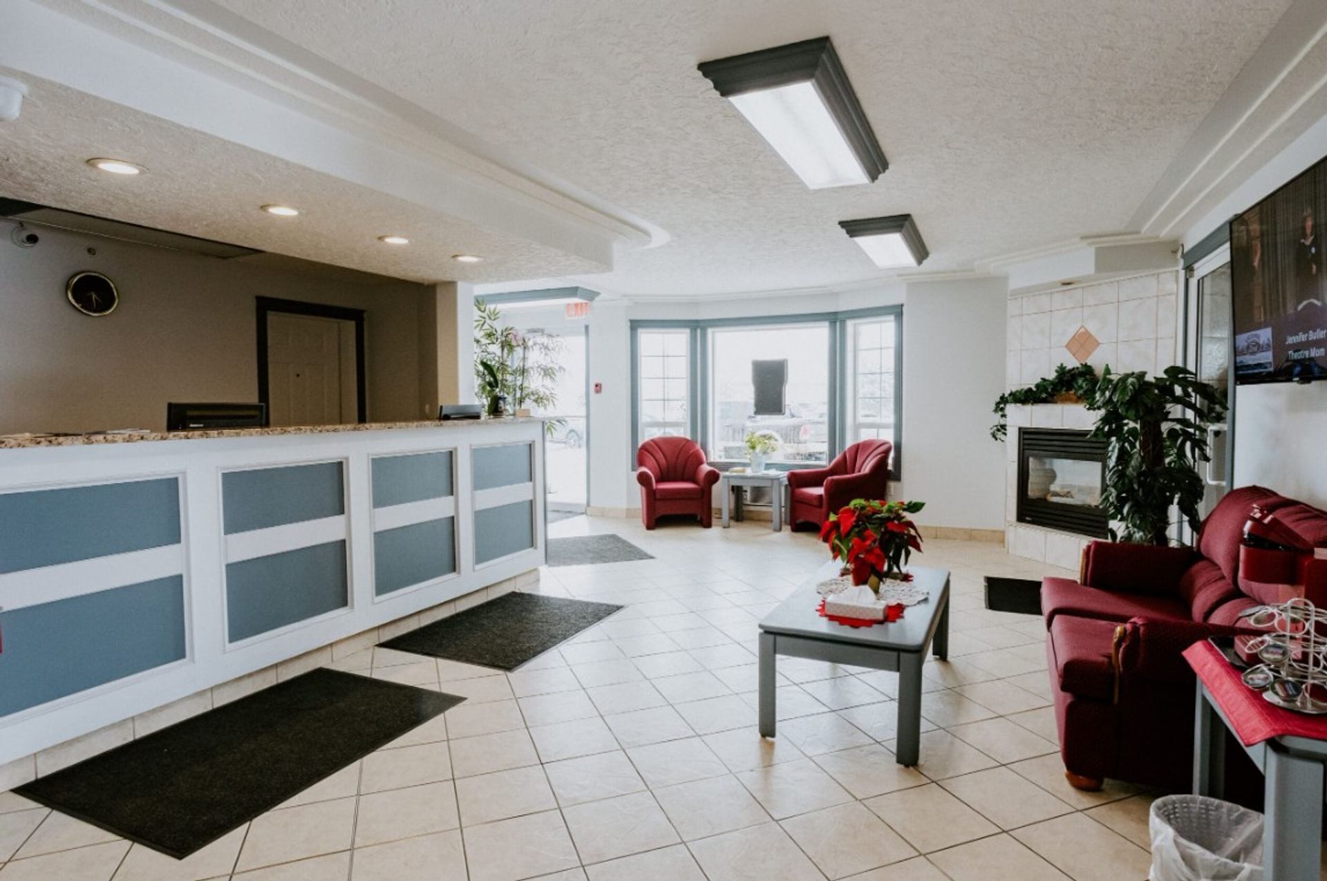 Hotel lobby with front desk, red armchairs, fireplace, plants and tiled floor.
