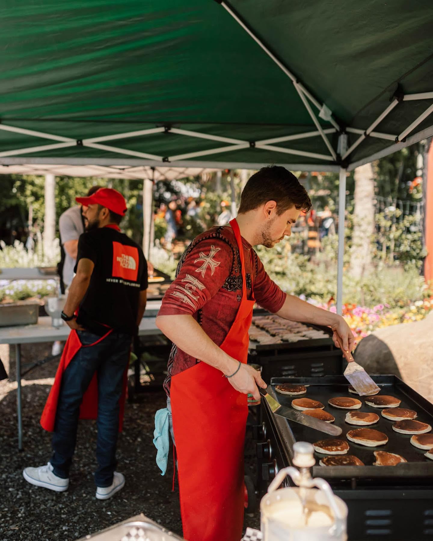 Cook in red apron flips pancakes on outdoor griddle under green canopy tent.