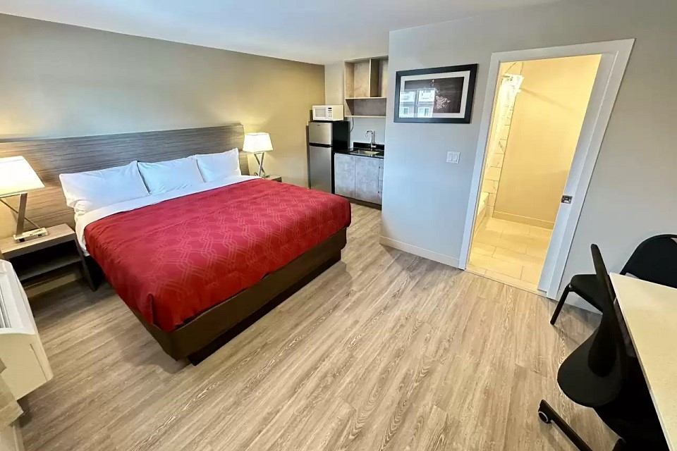 Hotel room with red bedspread, kitchenette, desk, and warm lighting.