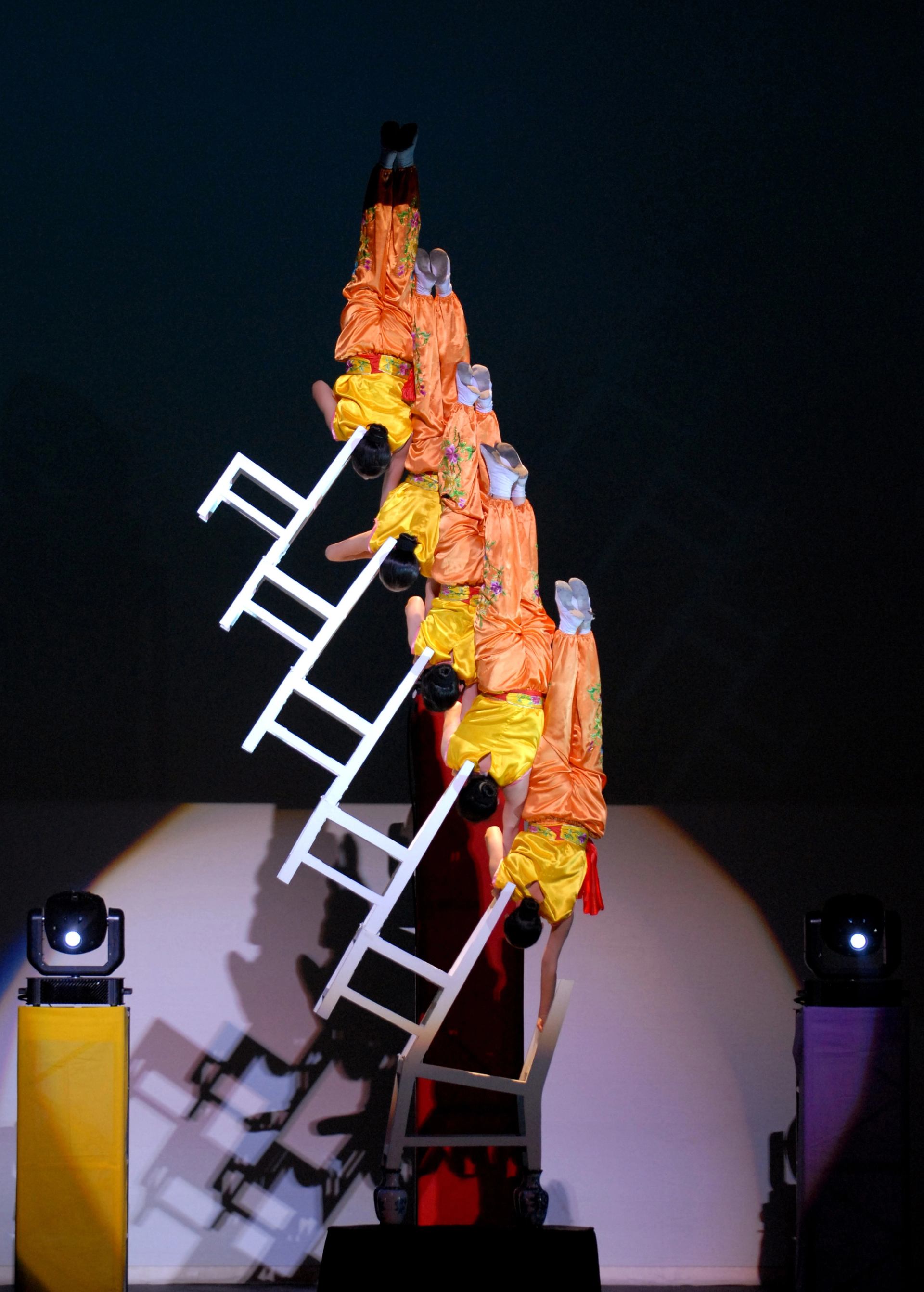 Acrobats in orange and yellow costumes balance upside down on stacked white ladders.