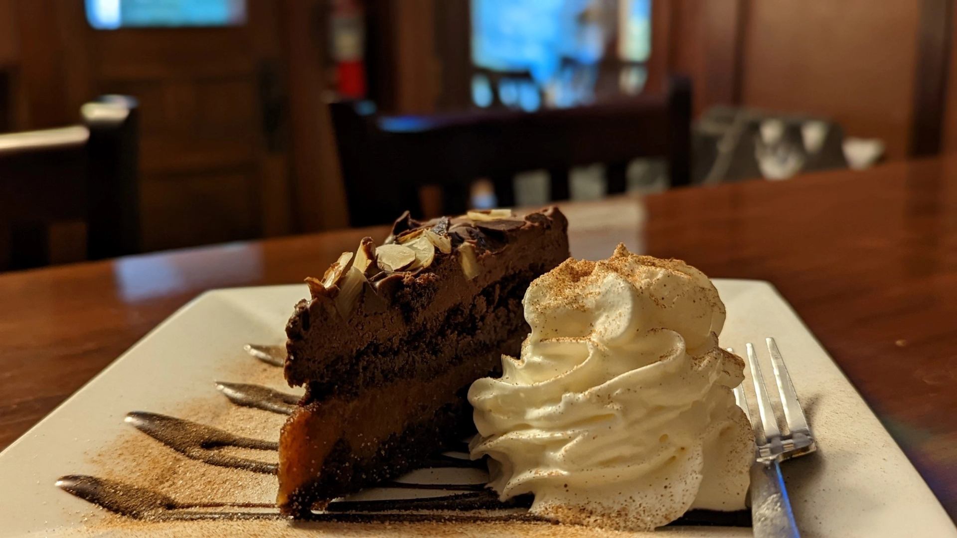 Chocolate cake slice with whipped cream and almonds served on a white plate with drizzle design.
