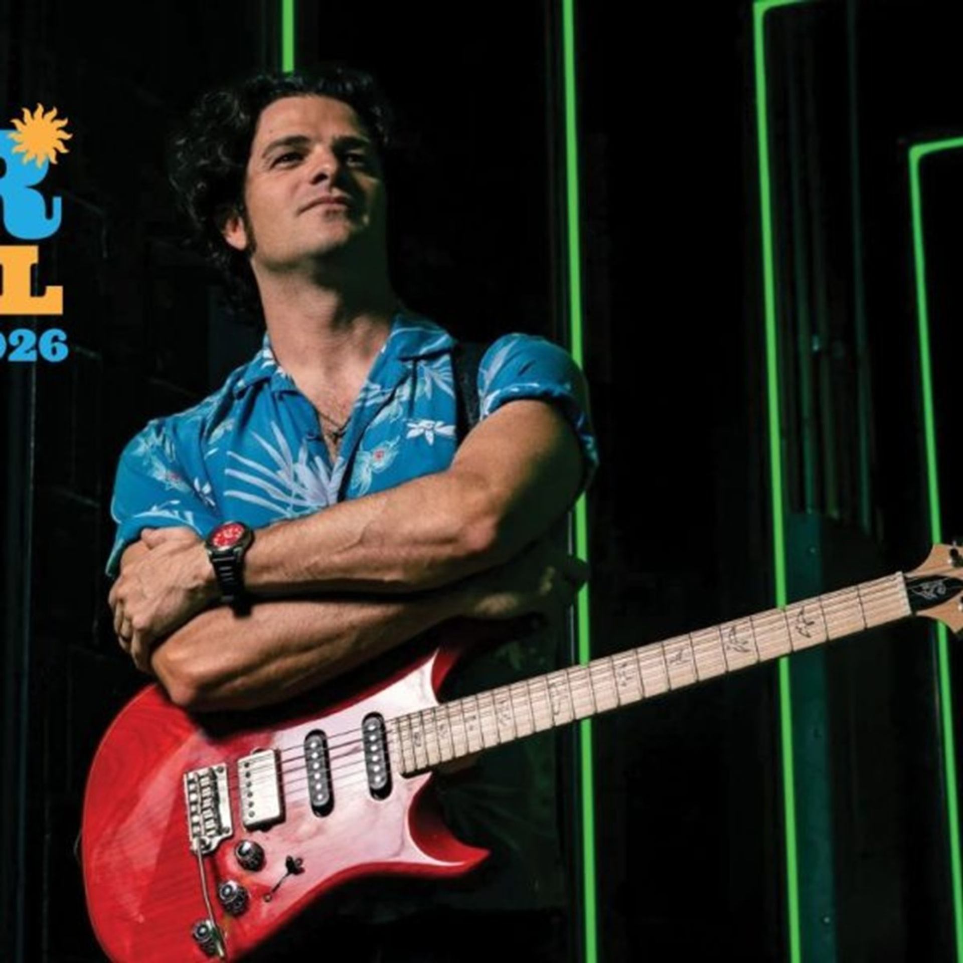 Musician in blue patterned shirt crosses arms while holding red electric guitar on stage.