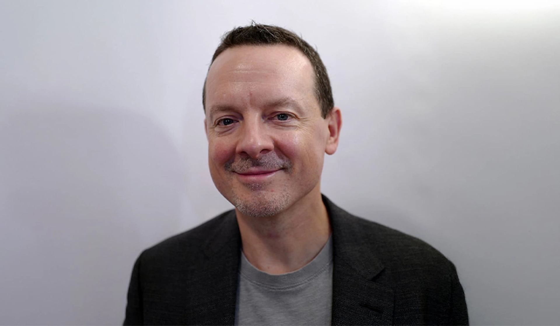 Person wearing a dark blazer and grey shirt against a plain light background.