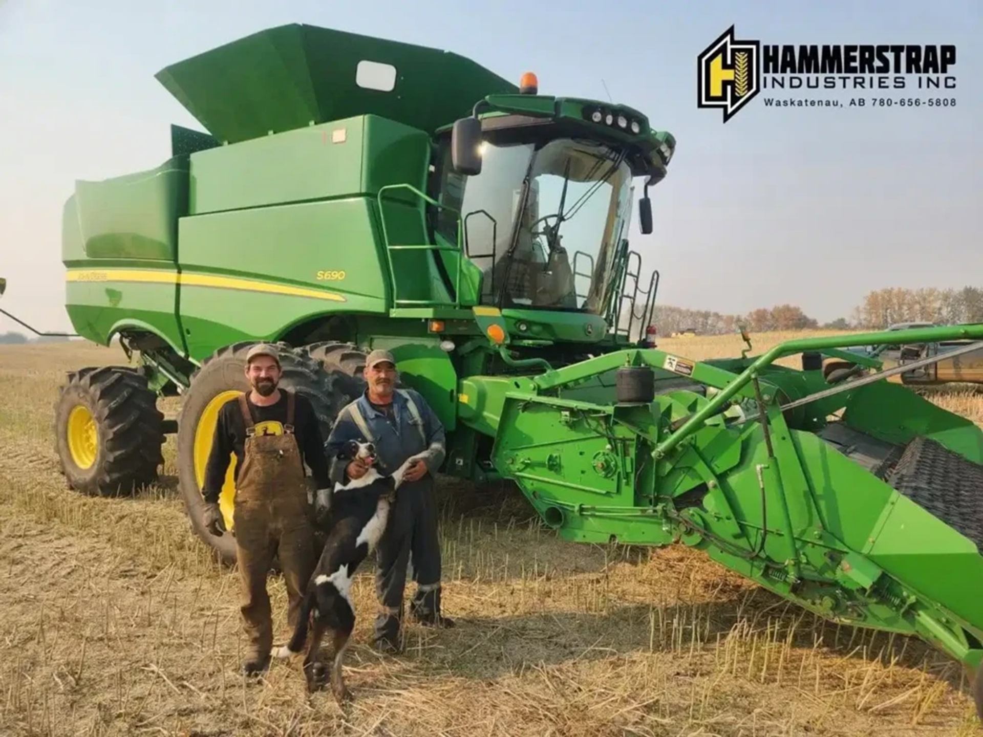 Farm workers and dog standing beside large green combine harvester in stubble field.