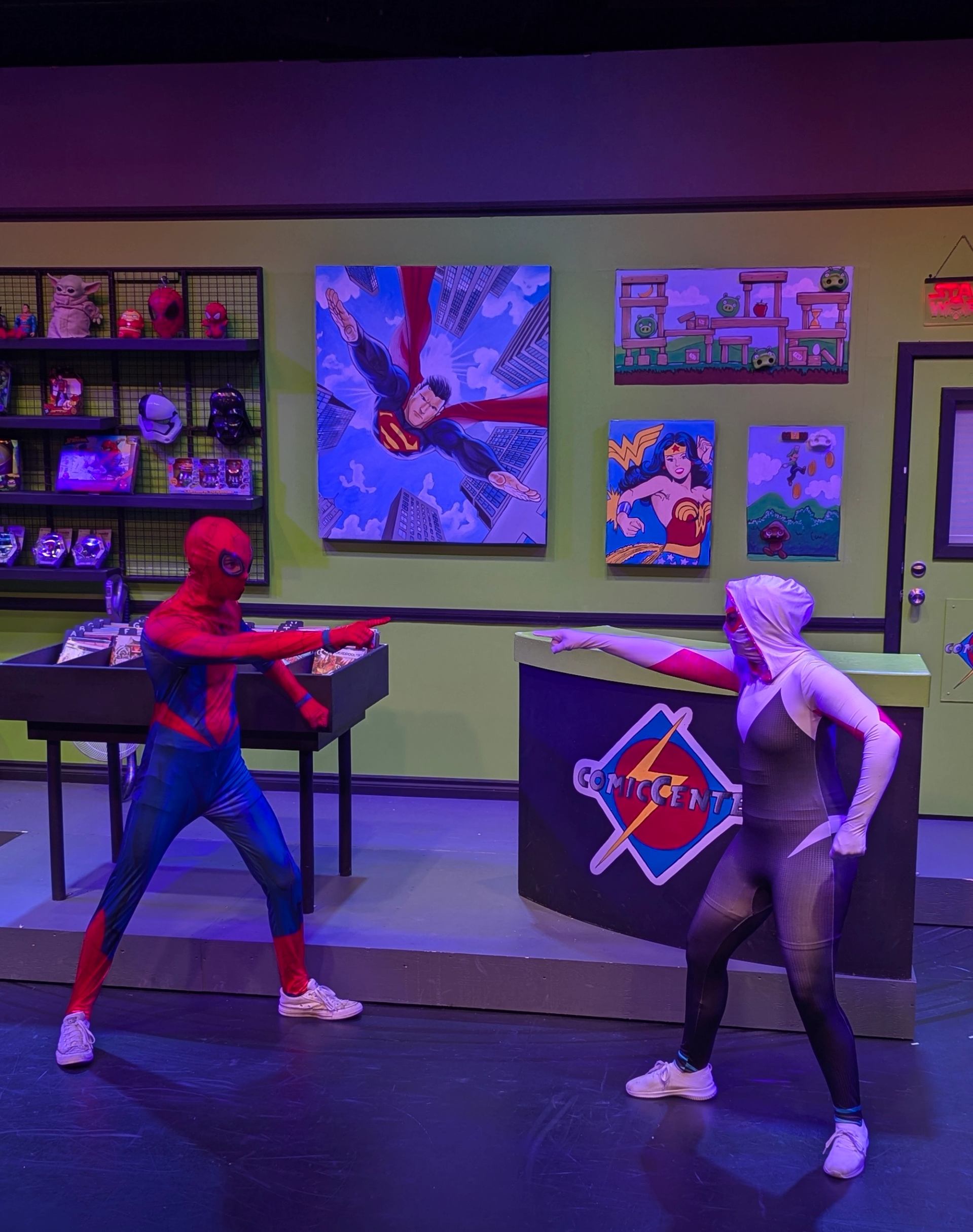 Comic-style costumed characters pose with painted backdrops inside a theatre venue.
