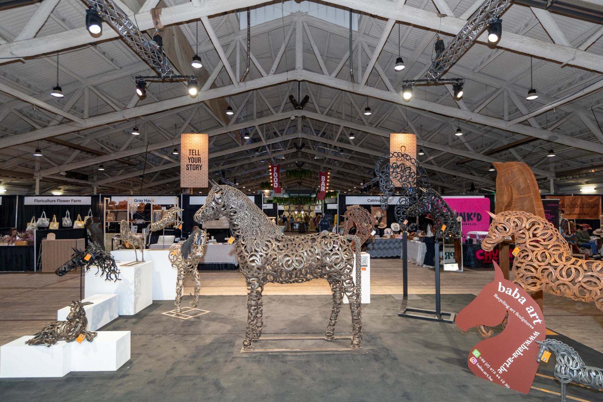 Indoor art exhibition at Spruce Meadows featuring metal horse sculptures, vendor booths, and overhead lighting.