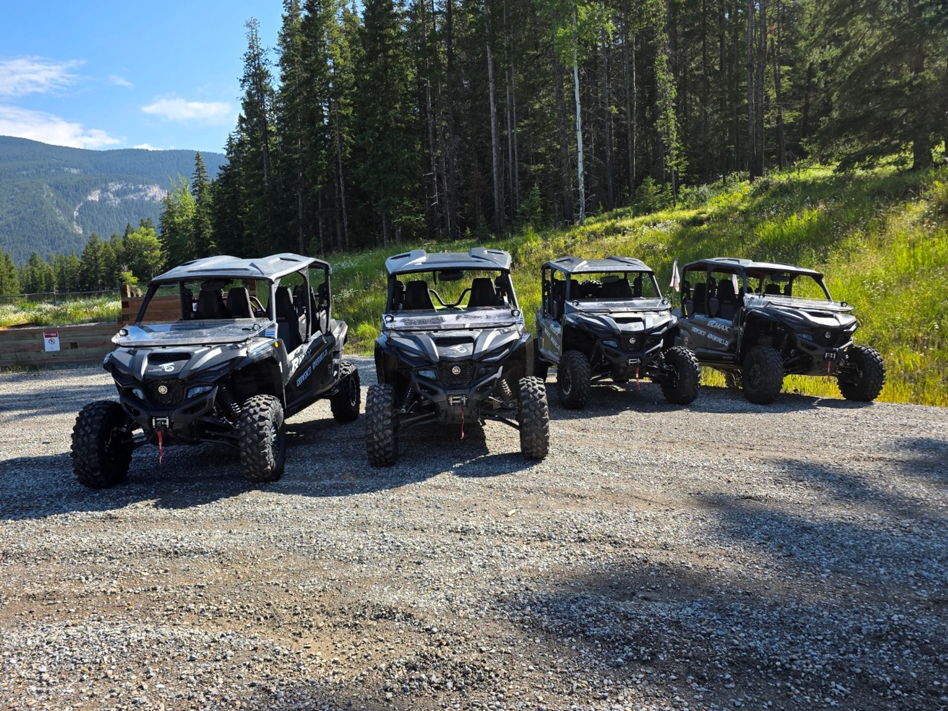Off-road side-by-side ATVs parked on gravel road beside a forest, with passengers visible.