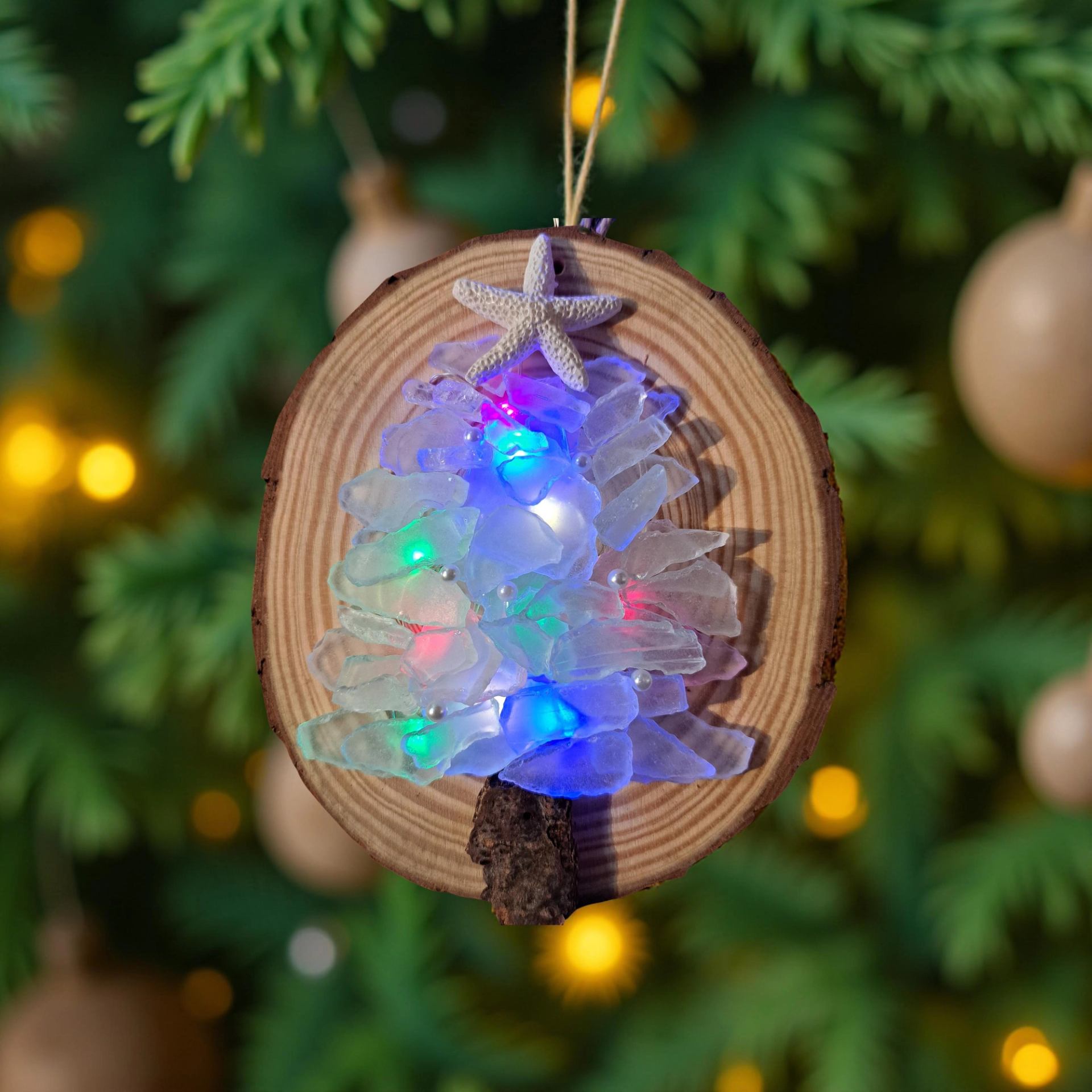 Wood slice ornament with sea glass tree and coloured lights hanging on evergreen.
