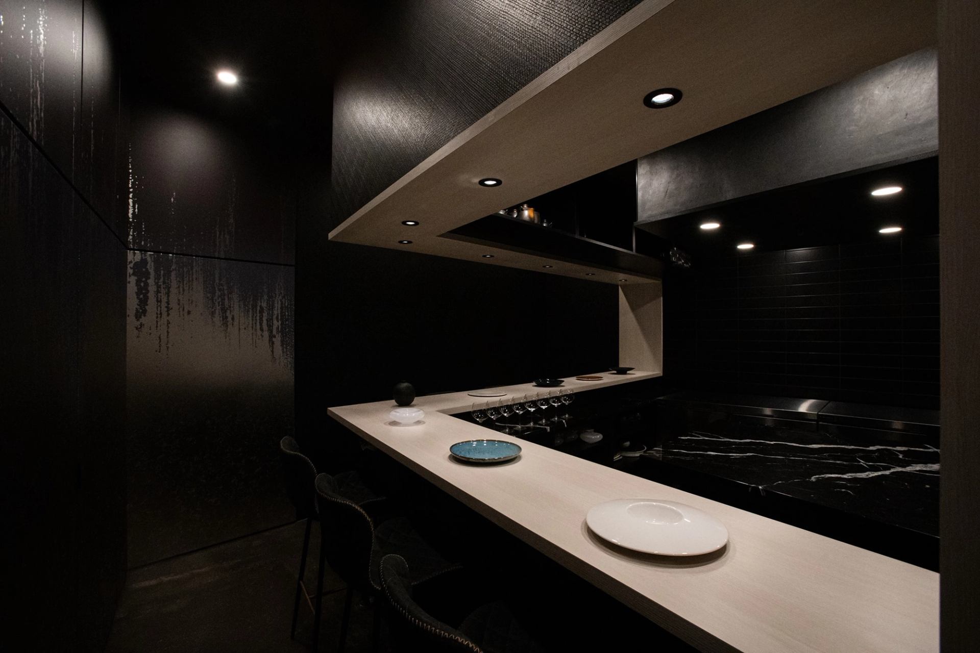 Dark modern kitchen with light countertop, bar stools, plates and recessed ceiling lights.