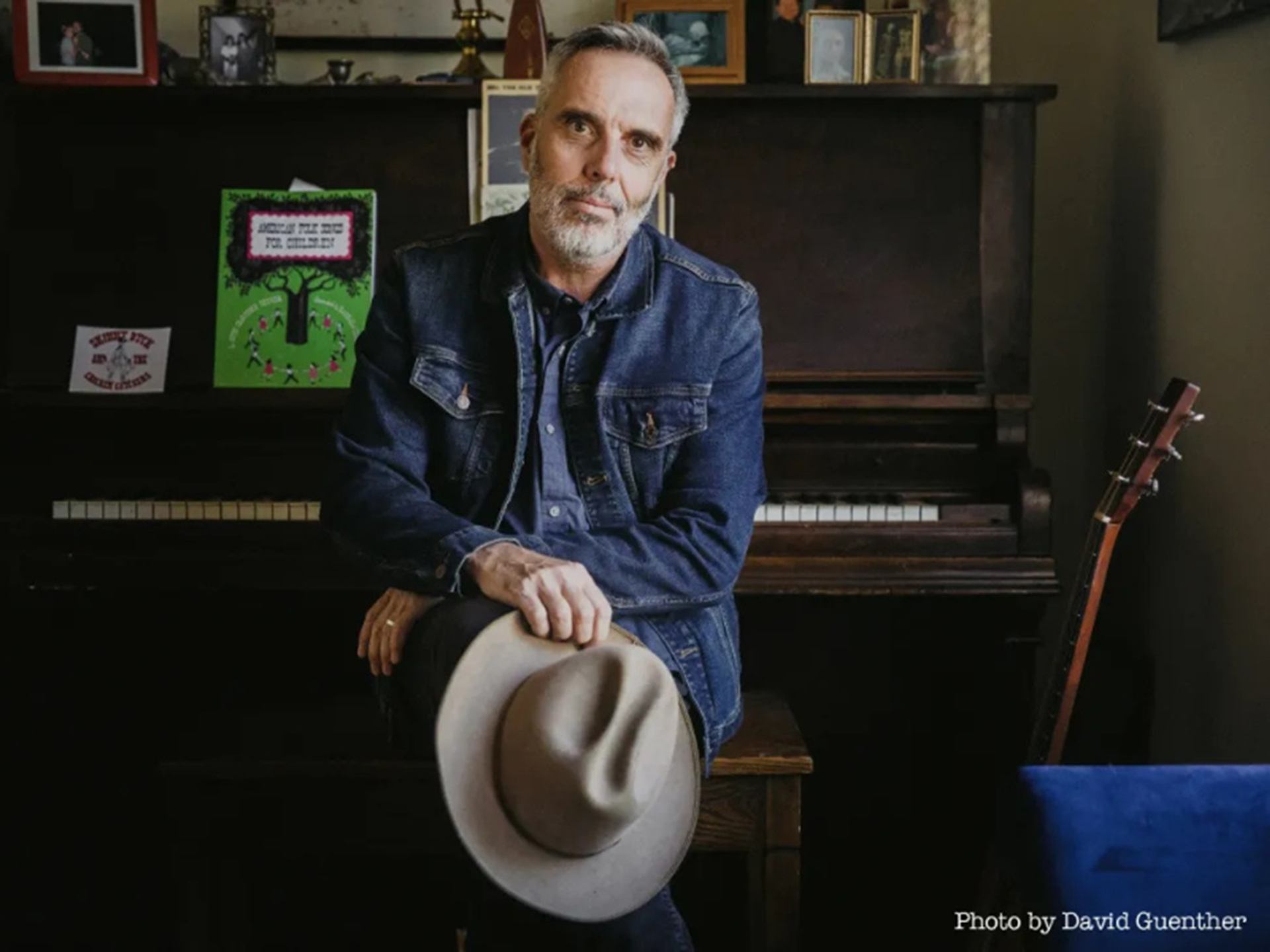 A man in a denim jacket sits on a piano bench, holding a hat, with an upright piano and guitar behind him.