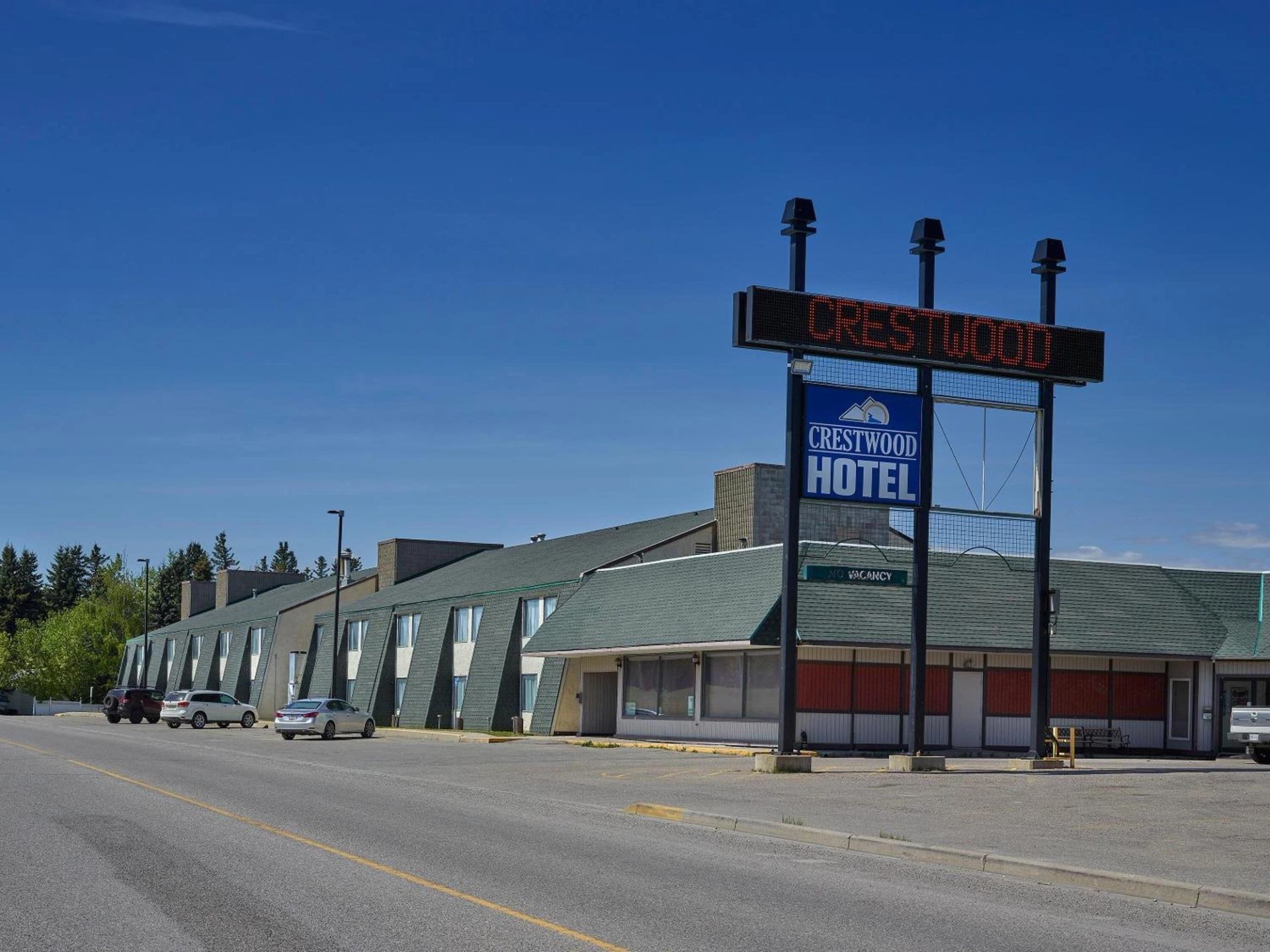 Exterior view of the Crestwood Hotel, a building with a green roof and slanted windows, next to its large roadside sign under a clear blue sky.