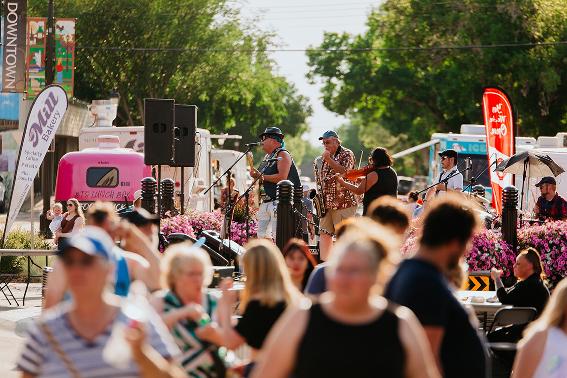 Outdoor band performs on street stage as crowd gathers near food trucks and flowers.