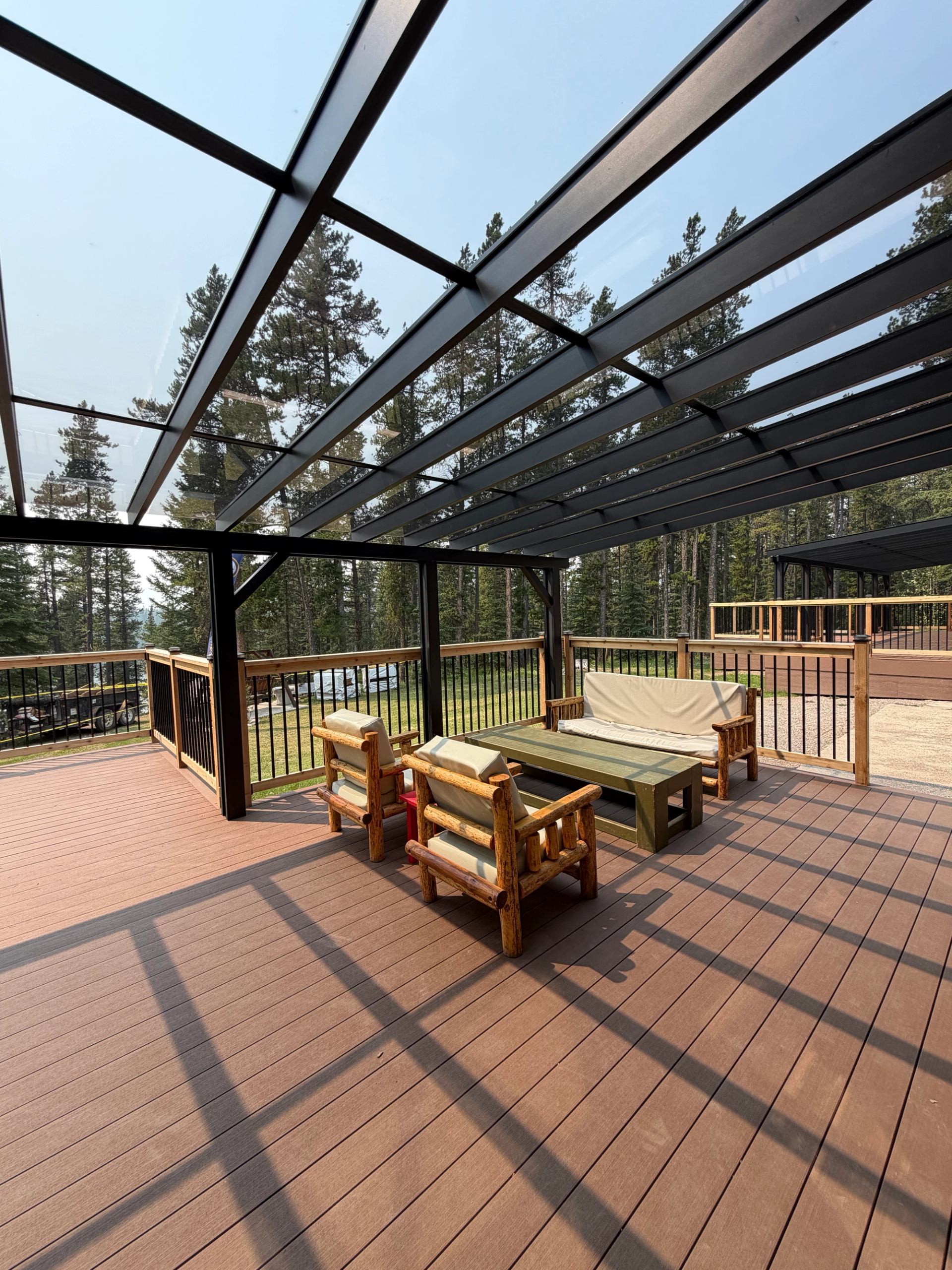 Covered wooden deck with log patio furniture under glass roof, overlooking tall evergreen trees.