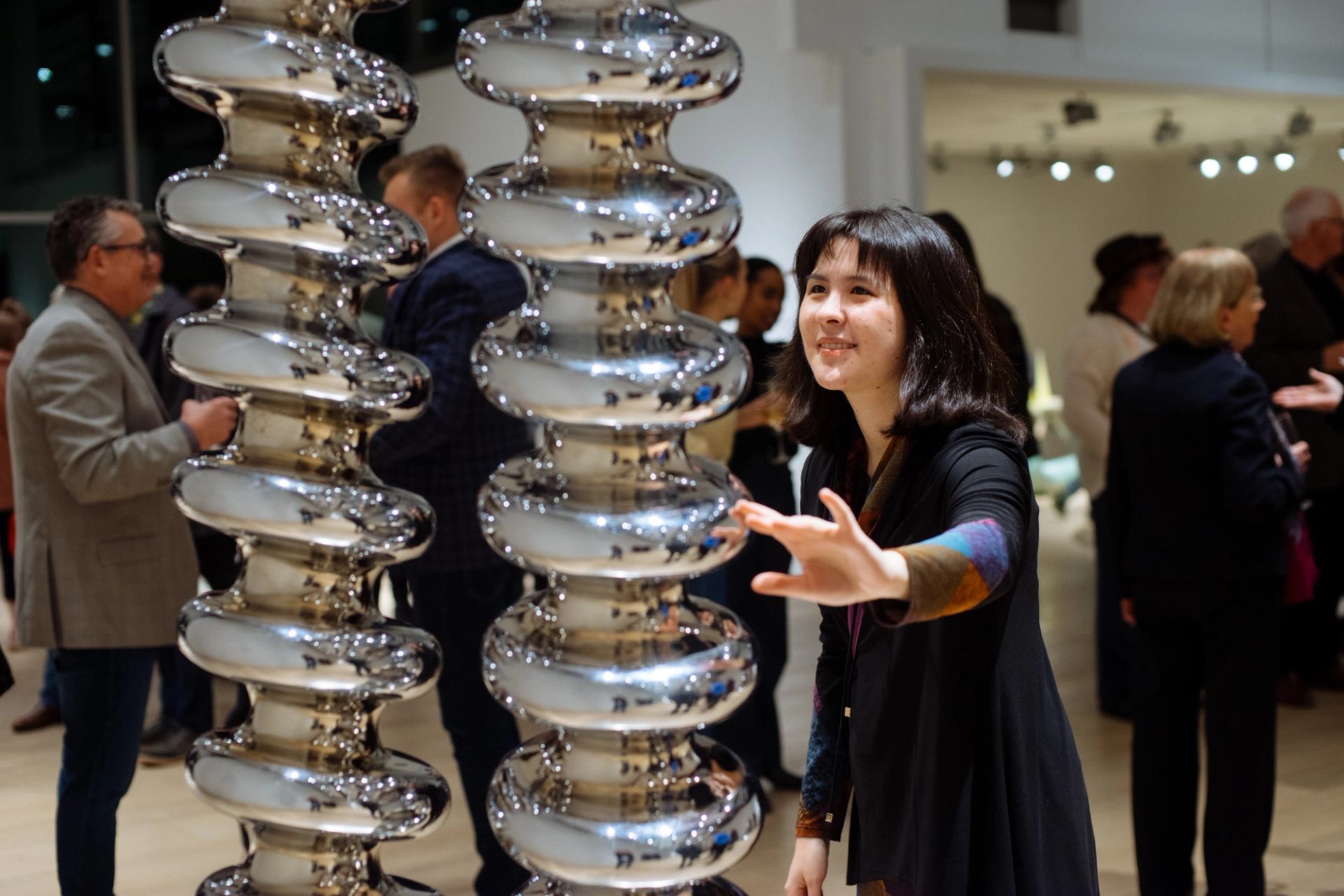 Woman interacting with shiny metallic sculpture at crowded art gallery opening, with people chatting around.