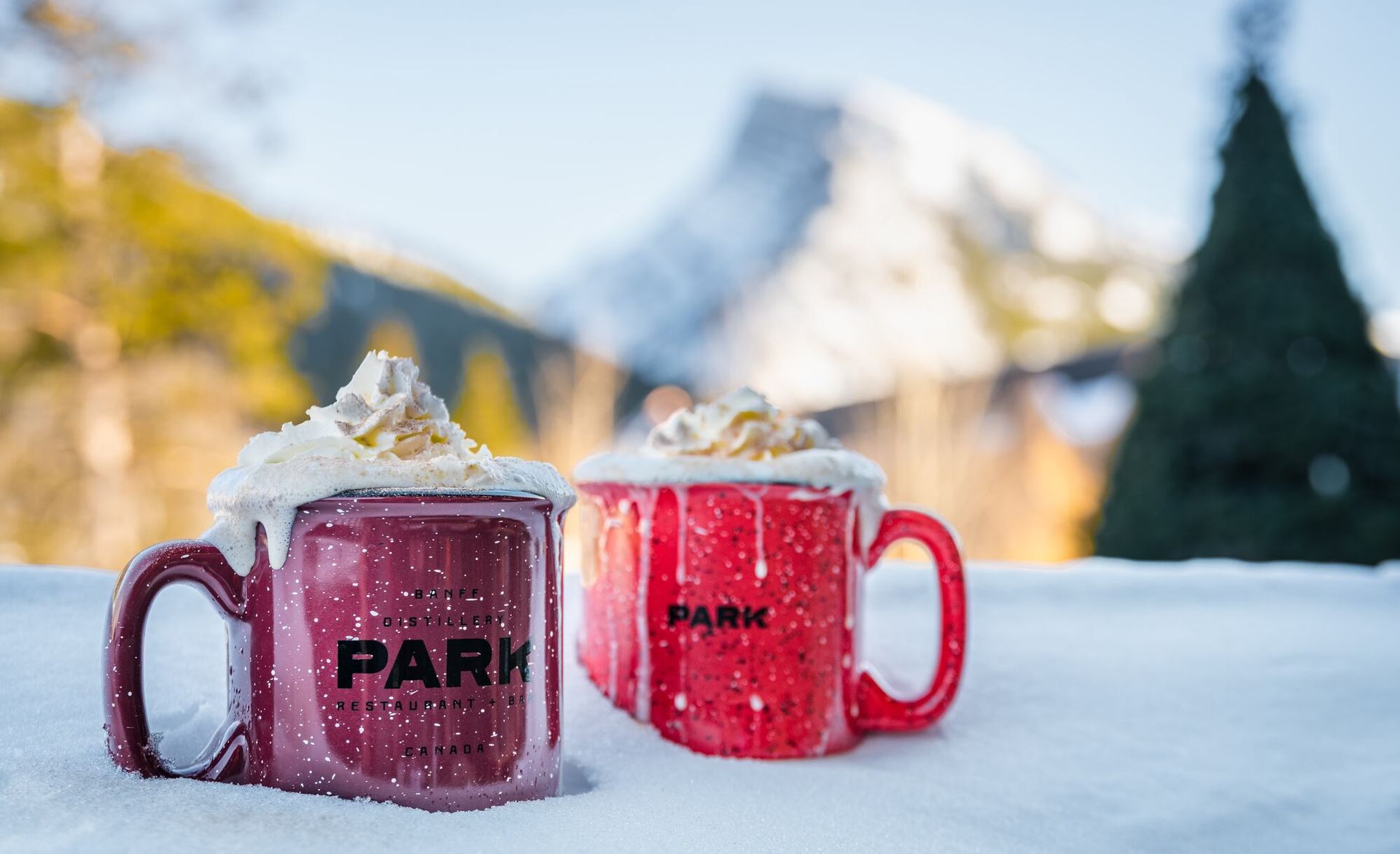 Two red mugs of hot chocolate with whipped cream and caramel on snowy mountain backdrop.