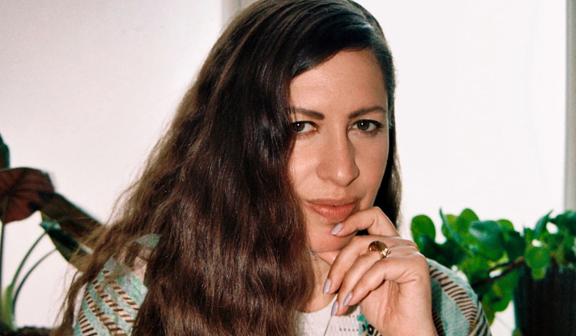 Person with long wavy hair rests chin on hand, indoor plants in background.